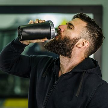 caesin protein powder front view portrait of young caucasian man athlete in black hoodie male standing in the gym holding protein supplement shaker supplementation in training waist up black hair and beard copy space drink